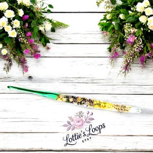 May include: A green and clear crochet hook with dried flowers and gold flakes embedded in the handle. The hook is lying on a white wooden surface with flowers on either side. The text "Lottie's Loops Etsy" is visible in the lower center of the image.