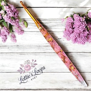 May include: A crochet hook with a gold-colored metal tip and a pink resin handle. The handle is decorated with gold leaf and floral elements. The hook is set against a white wooden background with purple flowers.