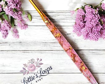 May include: A crochet hook with a gold-colored metal tip and a pink resin handle. The handle is decorated with gold leaf and floral elements. The hook is set against a white wooden background with purple flowers.