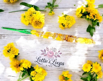 May include: A green and gold crochet hook with pressed flowers and gold leaf embedded in the resin. The hook is on a white wooden surface with yellow flowers scattered around it. The text "Lottie's Loops Etsy" is visible in the center of the image.