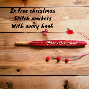May include: A red crochet hook with gold glitter and the words "Merry Christmas" written on it. The hook is lying on a wooden surface with red berries and stems.