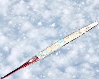 May include: A close-up of a silver and iridescent crochet hook with a red handle. The hook is lying on a white background with a snowflake pattern.