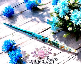 May include: A turquoise and gold resin crochet hook with a clear tip, resting on a light wood surface. The hook is decorated with blue and gold accents. The image also features blue flowers and the text "Lottie's Loops".