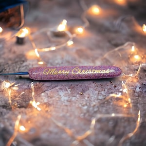 May include: A pink and purple glitter crochet hook with a gold inscription that reads "Merry Christmas".