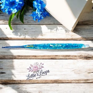 Può includere: Un uncinetto blu con un manico in resina trasparente riempito di glitter blu e verdi. L'uncinetto è su una superficie di legno con un logo floreale che dice "Lottie's Loops Etsy".