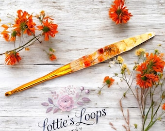 May include: A gold crochet hook with a clear handle filled with dried orange and yellow flowers. The hook is lying on a white wooden surface with orange flowers scattered around it. The text "Lottie's Loops Etsy" is visible in the lower center of the image.