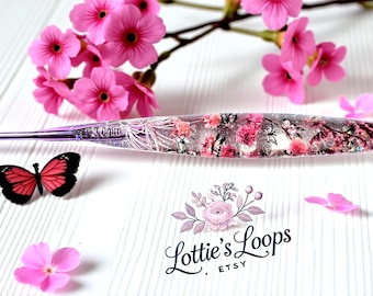 May include: A clear crochet hook with a floral design, featuring pink flowers and silver glitter. A small, red and black butterfly sits near the hook. The text "Lottie's Loops" is visible.