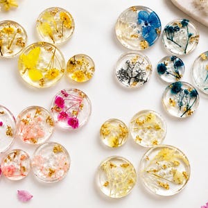 May include: A collection of circular resin cabochons featuring embedded dried flowers in various colors including yellow, pink, blue, and white, accented with small gold flakes throughout the clear, smooth, and glossy surfaces.