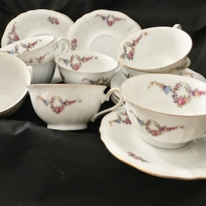 Könnte beinhalten: Ein Set aus sechs weißen Teetassen und Untertassen mit Blumendesigns in Rosa-, Blau- und Grüntönen. Die Tassen und Untertassen haben einen goldenen Rand.