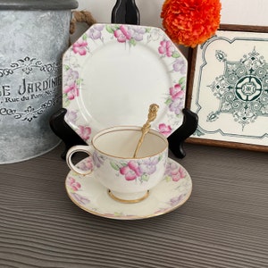 Könnte beinhalten: Eine weiße Teetasse und Untertasse mit einem Blumenmuster aus rosa und violetten Blumen. Die Untertasse hat einen goldenen Rand. Ein goldener Teelöffel befindet sich in der Teetasse.