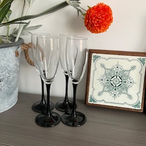 Op de afbeelding: Vier champagnefluiten met zwarte stelen, tegen een decoratieve tegel met een groen en wit ontwerp.