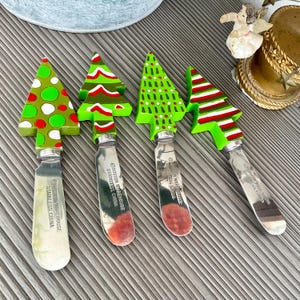 May include: Set of four stainless steel Christmas tree-shaped butter knives. Each knife has a different green, red, and white patterned design. The knives are made in China.