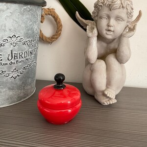 以下が含まれることがあります： 蓋に黒いノブが付いた、小さくて丸い赤いセラミックジャー。ジャーは、"JARDINIER"という文字が印刷された装飾的な金属製のバケツとケルビムの像の隣の灰色の表面に置かれています。