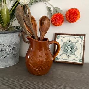 Pode incluir: Um jarro de cerâmica marrom com um design de pássaro contém utensílios de cozinha de madeira. O jarro está sobre uma superfície de madeira escura com um azulejo decorativo atrás.