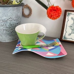 May include: A green and white ceramic teacup with a wavy handle sits on a colorful ceramic saucer with a fork. The saucer has a geometric pattern in shades of pink, blue, yellow, and white.