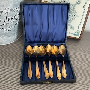 May include: A set of six gold-toned metal spoons arranged in a dark blue velvet-lined presentation box. The spoons feature a smooth, reflective finish and a tapered handle design. The box has a hinged lid with a deep blue satin interior lining.