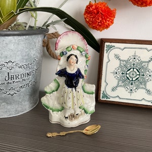 Op de afbeelding: Een porseleinen beeldje van een vrouw in een blauw-witte jurk met een groen-wit bloemenmotief. Het beeldje houdt een boeket bloemen vast en staat op een voet met een gouden rand. Het beeldje staat op een houten tafel met een groene en witte tegelonderzetter en een gouden lepel.