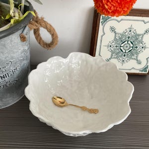 Peut inclure: Un bol en céramique blanche avec un bord texturé et plissé. Une cuillère à café en or repose à l'intérieur du bol.
