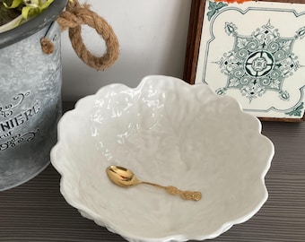 May include: A white ceramic bowl with a textured, ruffled edge. A gold teaspoon is resting inside the bowl.