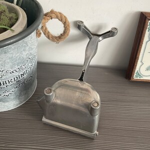 以下が含まれることがあります： ハンドルと3本の脚が付いたベースを持つ銀色の金属製のアイスクリームスプーン。スプーンは灰色の表面にあります。
