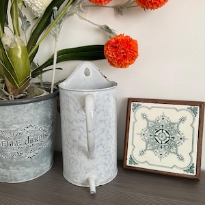 Puede incluir: Una regadera de metal blanco y azul con un patrón de mármol, con asa y boquilla. Una baldosa decorativa con un diseño floral verde y blanco en un marco de madera está junto a la regadera. Una planta en maceta y flores naranjas también están en la imagen.