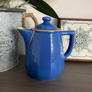 Op de afbeelding: Een kobaltblauwe keramische theepot met een gouden rand en handvat. De theepot heeft een geometrisch ontwerp en een gerepareerde scheur met gouden accenten. Een decoratief stuk voor de keuken of eetkamer.