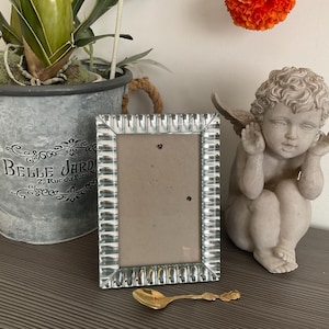 Op de afbeelding: Een rechthoekige zilveren fotolijst met een decoratieve rand. Een kleine gouden lepel ligt voor het frame. Een klein beeldje van een engel staat rechts van het frame. Een metalen plantenbak met een plant staat links.