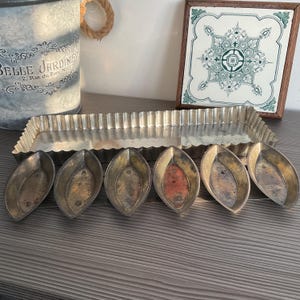 May include: A collection of vintage metal baking molds. Includes a rectangular fluted tart pan and six individual leaf-shaped molds. The metal has a tarnished, aged appearance, suggesting antique or vintage items. A decorative tile and a metal bucket are in the background.