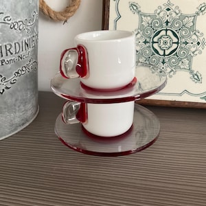 May include: Two stacked espresso cups and saucers made of white opaque glass. Each cup features a clear glass handle with a deep red accent. The saucers are clear glass with a distinct red rim, creating a layered appearance for the set.