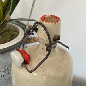 May include: A white ceramic bottle with a red rim and a metal wire closure. The bottle has a rustic, vintage look.