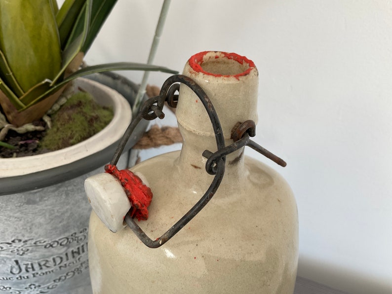 May include: A white ceramic bottle with a red rim and a metal wire closure. The bottle has a rustic, vintage look.