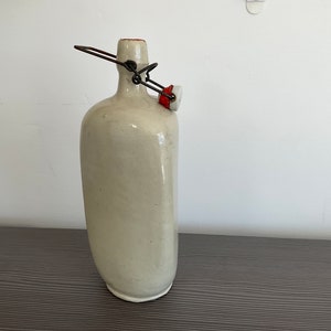 May include: A white ceramic bottle with a metal and red stopper. The bottle has a rounded shape and a smooth surface.
