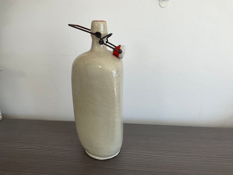 May include: A white ceramic bottle with a metal and red stopper. The bottle has a rounded shape and a smooth surface.