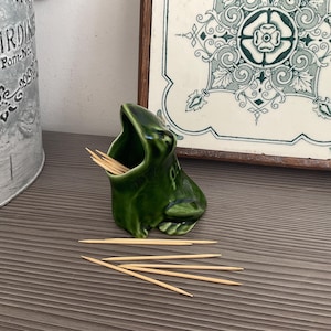 May include: A green ceramic frog-shaped toothpick holder with a wide open mouth, filled with wooden toothpicks. Several toothpicks are scattered in front of the frog. A decorative floral tile is in the background.