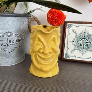 Può includere: Una tazza tiki in ceramica gialla con un design a forma di faccia sorridente. La tazza è accanto a una fioriera in metallo zincato con le parole "Belle Jardiniere" e una piastrella decorativa con un motivo verde e bianco.