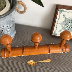 May include: A vintage wooden coat rack with three rounded knobs. The rack is a warm orange color and has a bamboo-like design. A small gold spoon rests below the rack. A decorative tile and a metal pot are in the background.
