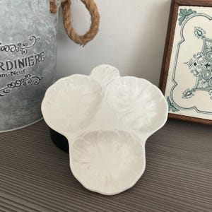 Puede incluir: Una fuente blanca de tres secciones con un diseño en forma de hoja. La fuente está hecha de cerámica y tiene un acabado brillante. La fuente está diseñada para servir aperitivos o condimentos.