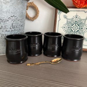 Może przedstawiać: Cztery małe, ciemnobrązowe ceramiczne garnuszki, każdy z wytłoczonym słowem "Deluxe" z przodu. Naczynia mają gładki, zaokrąglony kształt z lekko rozszerzanym brzegiem. Z przodu leży pojedyncza, złota metalowa łyżeczka z ozdobnym uchwytem.