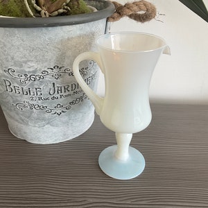 Puede incluir: Una jarra de vidrio opaco blanco con asa y base azul claro. La jarra está sobre una superficie de madera.