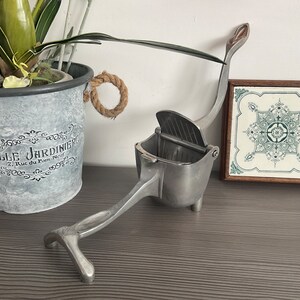以下が含まれることがあります： レバーとヒンジ付きの蓋が付いた銀色の手動柑橘類ジューサー。ジューサーは、緑と白のデザインの装飾タイルの隣にある木製の表面にあります。
