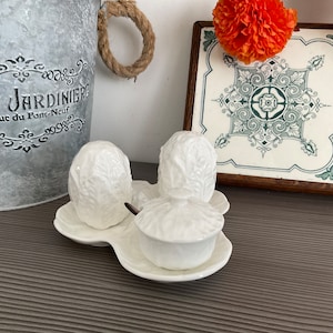 Pode incluir: Conjunto de saleiro e pimenteiro em cerâmica branca e uma pequena tigela com tampa numa bandeja a condizer. O conjunto tem um design texturizado semelhante a uma couve. Inclui uma colher pequena. Uma telha decorativa com um padrão floral no fundo.