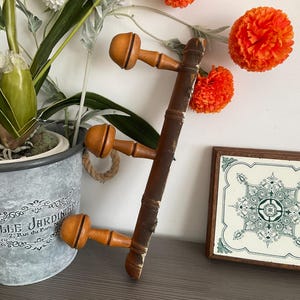 May include: A vintage wooden coat rack with three pegs, featuring a dark brown finish and light orange peg heads. The rack is displayed with a decorative tile and a potted plant, creating a rustic aesthetic.