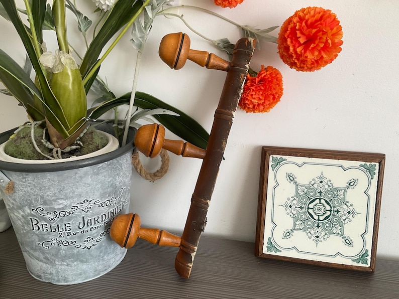 May include: A vintage wooden coat rack with three pegs, featuring a dark brown finish and light orange peg heads. The rack is displayed with a decorative tile and a potted plant, creating a rustic aesthetic.