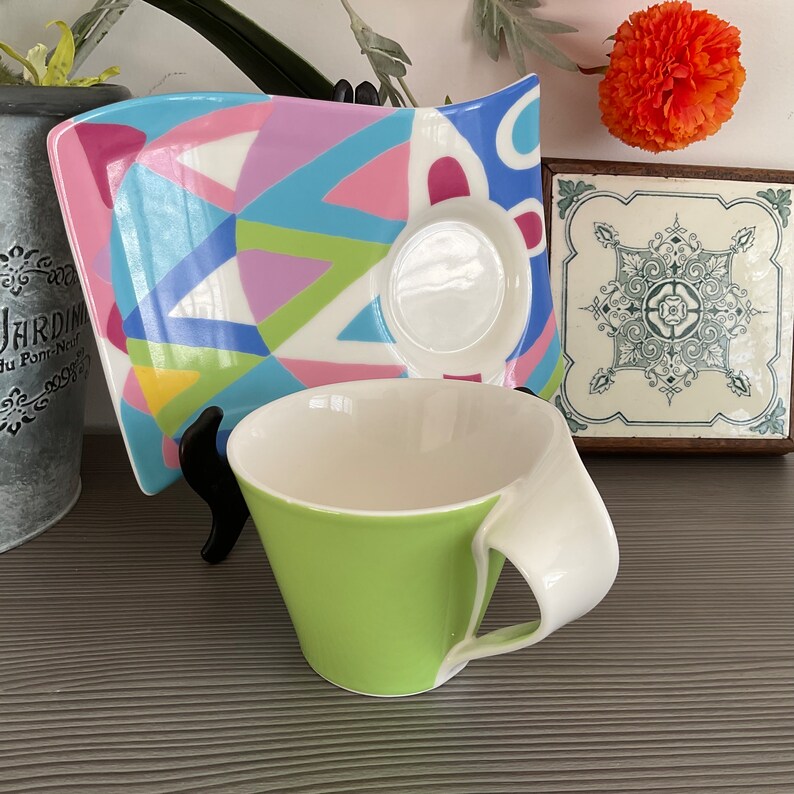 May include: A green and white ceramic teacup with a handle. The cup is sitting on a colorful, geometric patterned saucer. The saucer is on a wooden surface.