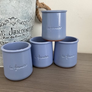 May include: Four pale blue ceramic jars, each with the embossed text "la fermière". The jars are cylindrical with a flared rim. A decorative metal bucket with text is in the background.