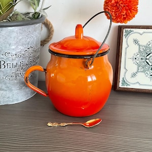 Puede incluir: Olla de esmalte naranja con tapa y asa de alambre. La olla tiene un degradado de colores naranja y rojo. La olla está sobre una superficie de madera.