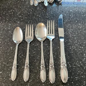 Könnte beinhalten: Ein Satz antiker Silberwaren, darunter ein Löffel, zwei Gabeln, ein Servierlöffel und ein Messer. Das Besteck hat ein Blumendesign auf den Griffen und eine polierte, reflektierende Oberfläche. Die Anordnung befindet sich auf einer dunklen, gesprenkelten Oberfläche.