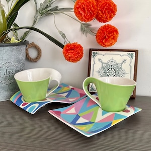 May include: Two green teacups with white interiors and matching saucers with a colorful geometric pattern. The saucers are connected together.