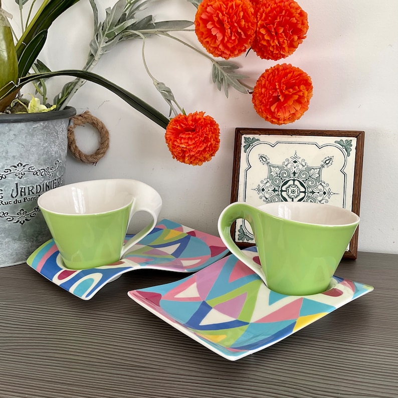 May include: Two green teacups with white interiors and matching saucers with a colorful geometric pattern. The saucers are connected together.