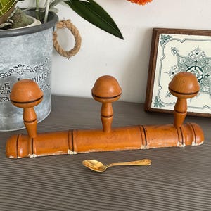 May include: An antique wooden coat rack with three rounded pegs, painted in a warm orange hue. A small gold spoon rests below. A decorative tile and a metal planter with a plant are in the background.